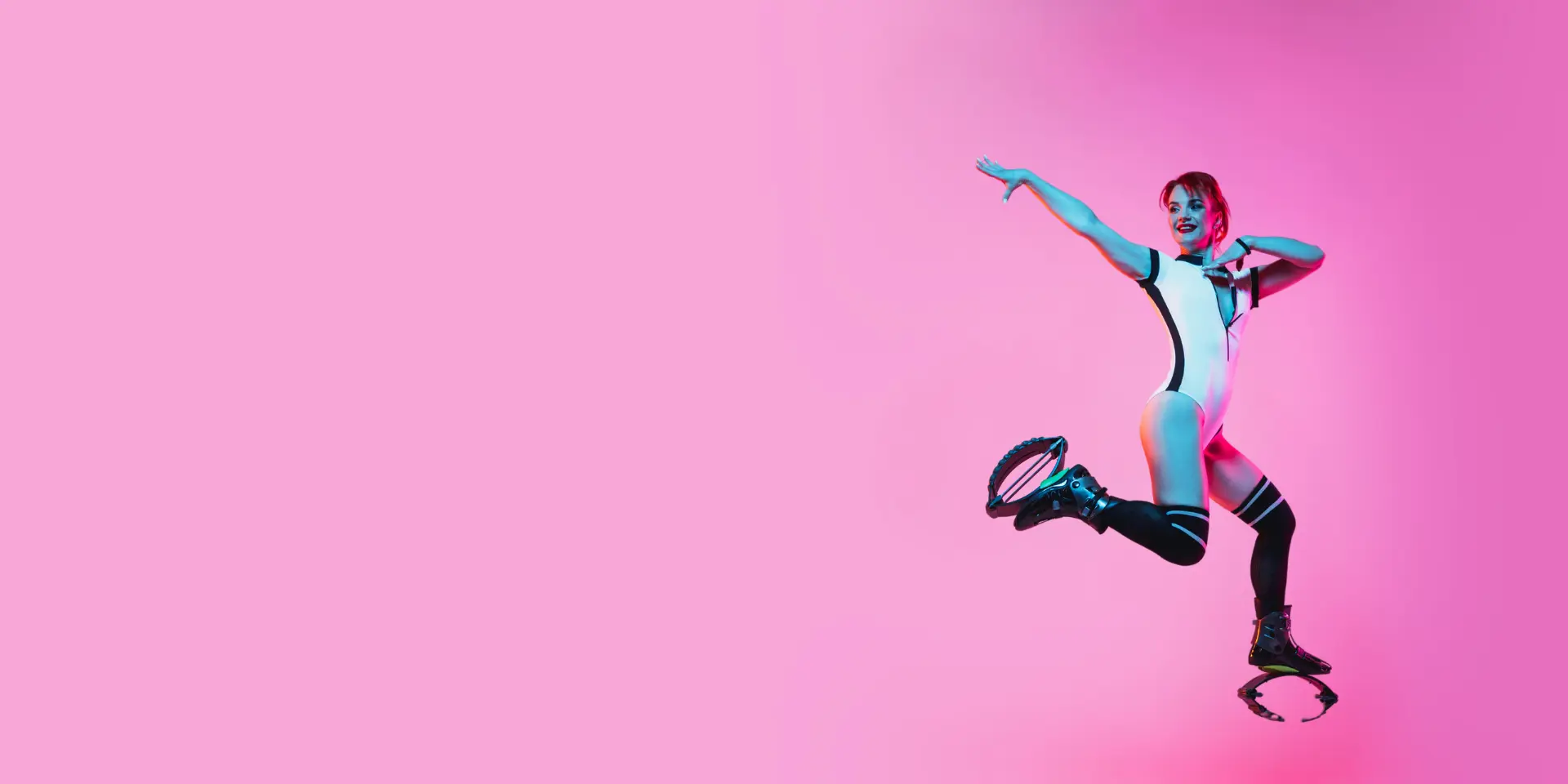 A woman in a dynamic mid-air pose wearing Kangoo Jumps rebound boots. She is dressed in a stylish white and black outfit, set against a vibrant pink background, showcasing energy and fitness. Kangojump dubai