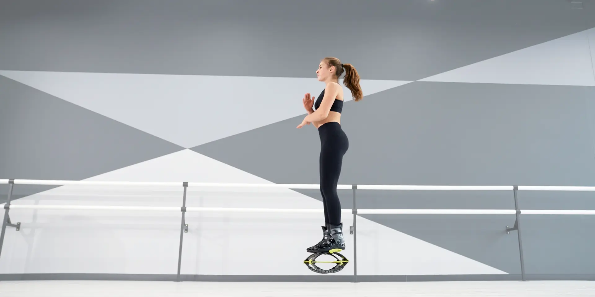 A woman performing a balanced pose on Kangoo Jumps rebound boots in a modern studio with geometric wall designs. She is dressed in black workout attire, highlighting focus and stability. Kangoo jump dubai