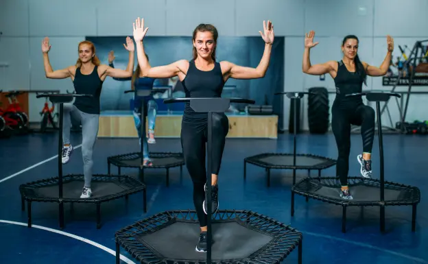 A group fitness class featuring participants on mini trampolines. They are engaged in synchronized aerobic exercises, raising their arms in an indoor gym setting with fitness equipment visible in the background. Kangoo Jump Dubai